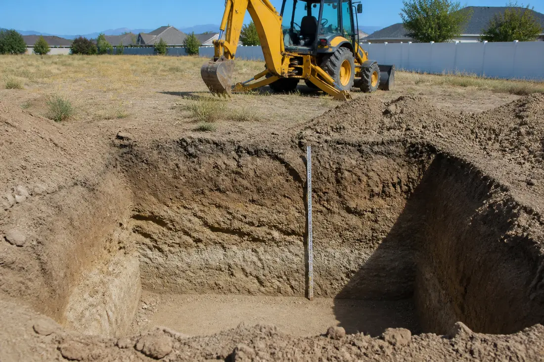 An open backhoe test pit on a residential lot with the soil profile visible in the wall